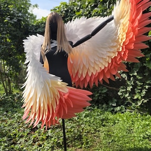 May include: Large decorative wings featuring a layered feather design that transitions in color from white at the top to a soft yellow in the middle and a vibrant coral pink at the tips. The wings are mounted on a black mannequin torso stand.