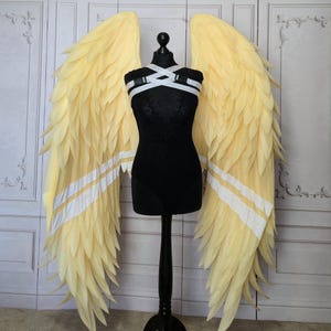 Puede incluir: Un par de alas de ángel amarillas con rayas blancas, exhibidas en un maniquí negro. Las alas están hechas de material plumoso en capas. El maniquí está sobre un soporte negro.