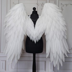 Könnte beinhalten: Große, weiße Engelsflügel mit einem geschichteten Federdesign. Die Flügel werden auf einer schwarzen Schaufensterpuppe präsentiert, die ihre volle Spannweite zeigt. Die Flügel bestehen aus einem strukturierten Material, das einen weichen, ätherischen Look erzeugt. Ideal für Kostüme oder dekorative Ausstellungen.