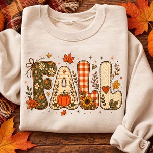 May include: A cream-coloured crewneck sweatshirt featuring the word FALL in large, decorative letters. Each letter has a unique pattern, including floral prints, orange gingham, and cable knit textures, accented with pumpkins and leaves.