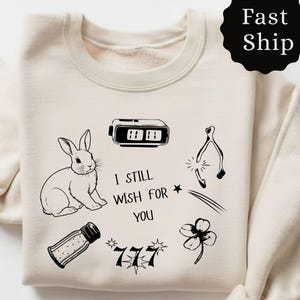 Op de afbeelding: Crèmekleurige sweatshirt met zwarte grafische illustraties, waaronder een konijn, wekker, wensbeen, klavertjevier, zoutstrooier en de tekst "I STILL WISH FOR YOU" en "777". Een zwarte cirkel met "Fast Ship" staat rechtsboven.