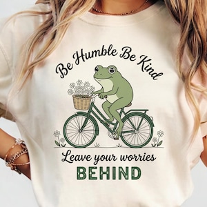 Pode incluir: Uma t-shirt cor de creme com um gráfico de um sapo verde a andar de bicicleta com um cesto de flores brancas. O texto diz Be Humble Be Kind e Leave your worries BEHIND em letras pretas e verdes com pequenos detalhes florais.
