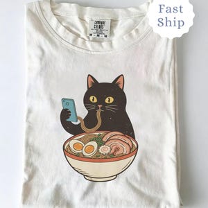 Op de afbeelding: Een witte keramische mok met een zwarte handgreep en de tekst "But First, Coffee" in een vetgedrukte, zwarte letter. De mok staat tegen een wazige achtergrond, wat het strakke ontwerp benadrukt.