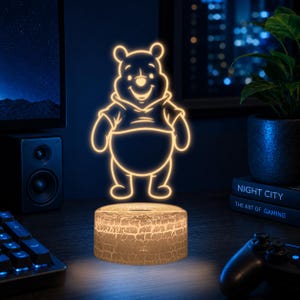May include: A 3D LED lamp featuring a glowing yellow outline of Winnie the Pooh. The light is emitted from a circular base with a textured, cracked white surface pattern. The character is depicted in a standing position with arms at the sides.