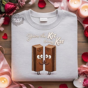 May include: A light gray sweatshirt with the embroidered phrase "You're the Kit to my Kat" and a cartoon illustration of two Kit Kat candy bars holding hands. The sweatshirt has a crew neck and long sleeves.