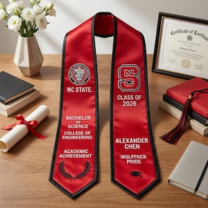May include: A red graduation stole with black trim, featuring the NC State logo and text. The stole includes the graduate's name, degree, and class year. A diploma, books, and flowers are in the background.