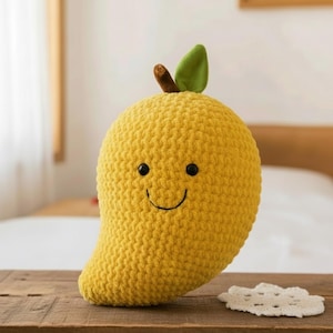 Puede incluir: Un peluche con forma de mango amarillo sonriente, con una hoja verde y un tallo marrón. El peluche tiene ojos negros y una sonrisa cosida. El juguete está sobre una superficie de madera.