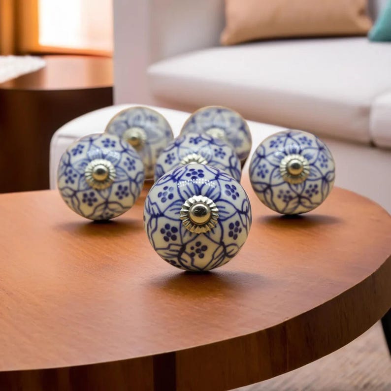 May include: A collection of six ceramic cabinet knobs with a blue and white floral pattern. Each knob has a brass-colored metal base. The knobs are arranged on a round, wooden table.