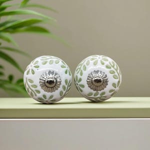 Op de afbeelding: Twee witte keramische kastknoppen met een bloemig bladerpatroon in lichtgroen. Elke knop heeft een zilverkleurige metalen basis en een centrale schroef. De knoppen zijn rond en liggen op een lichtgroen oppervlak.