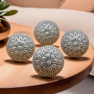 Peut inclure: Quatre boutons de meuble en c&eacute;ramique grise avec un motif floral. Les boutons sont ronds et pr&eacute;sentent un motif en relief textur&eacute;. Ils sont dispos&eacute;s sur une surface en bois.