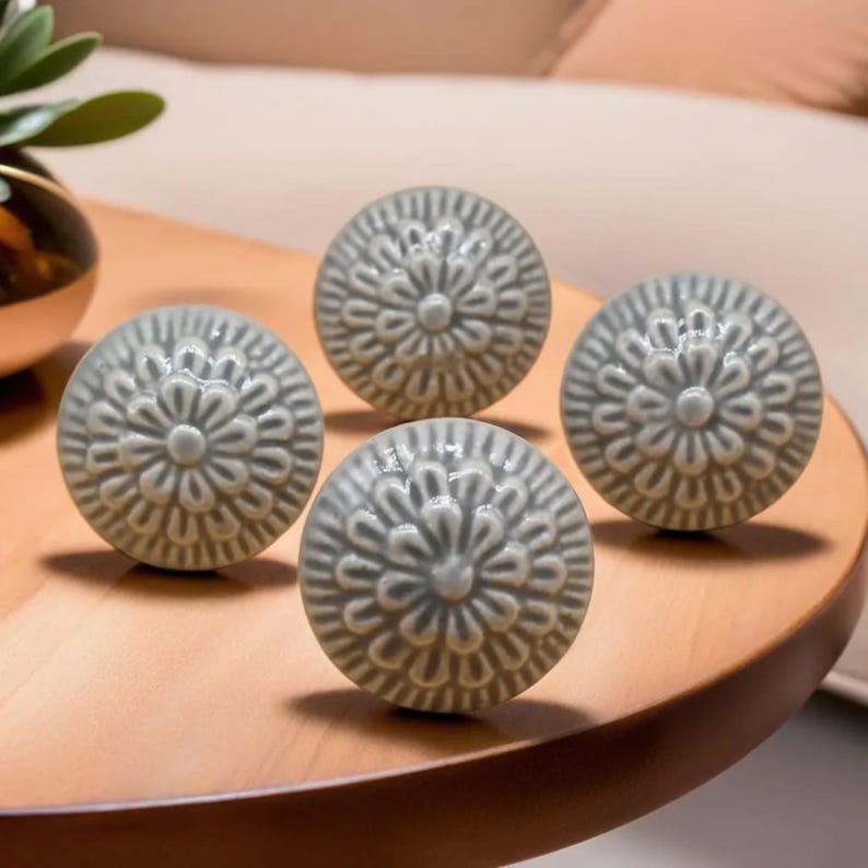 Peut inclure: Quatre boutons de meuble en c&eacute;ramique grise avec un motif floral. Les boutons sont ronds et pr&eacute;sentent un motif en relief textur&eacute;. Ils sont dispos&eacute;s sur une surface en bois.