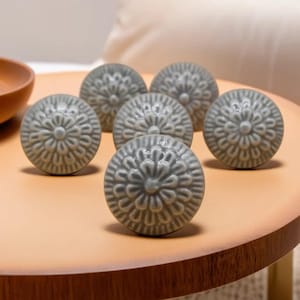 Peut inclure: Six boutons de meuble en céramique grise avec un motif floral sont disposés sur une surface brun clair. Les boutons sont ronds et présentent un motif en relief détaillé. Les boutons sont de couleur grise sourde.