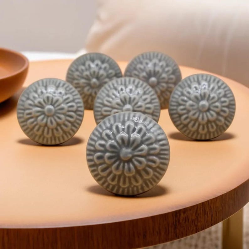 Peut inclure: Six boutons de meuble en c&eacute;ramique grise avec un motif floral sont dispos&eacute;s sur une surface brun clair. Les boutons sont ronds et pr&eacute;sentent un motif en relief d&eacute;taill&eacute;. Les boutons sont de couleur grise sourde.