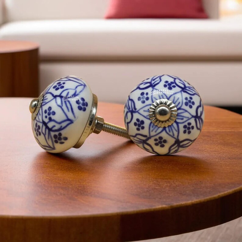 May include: Two decorative ceramic cabinet knobs. Each knob is round with a white base and a blue floral pattern. The knobs have metal hardware and are displayed on a wooden surface.