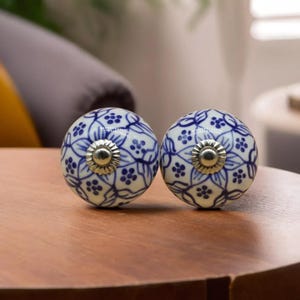 May include: Two round, white ceramic cabinet knobs with a blue floral pattern. Each knob has a silver-colored metal base. The knobs are on a wooden surface.
