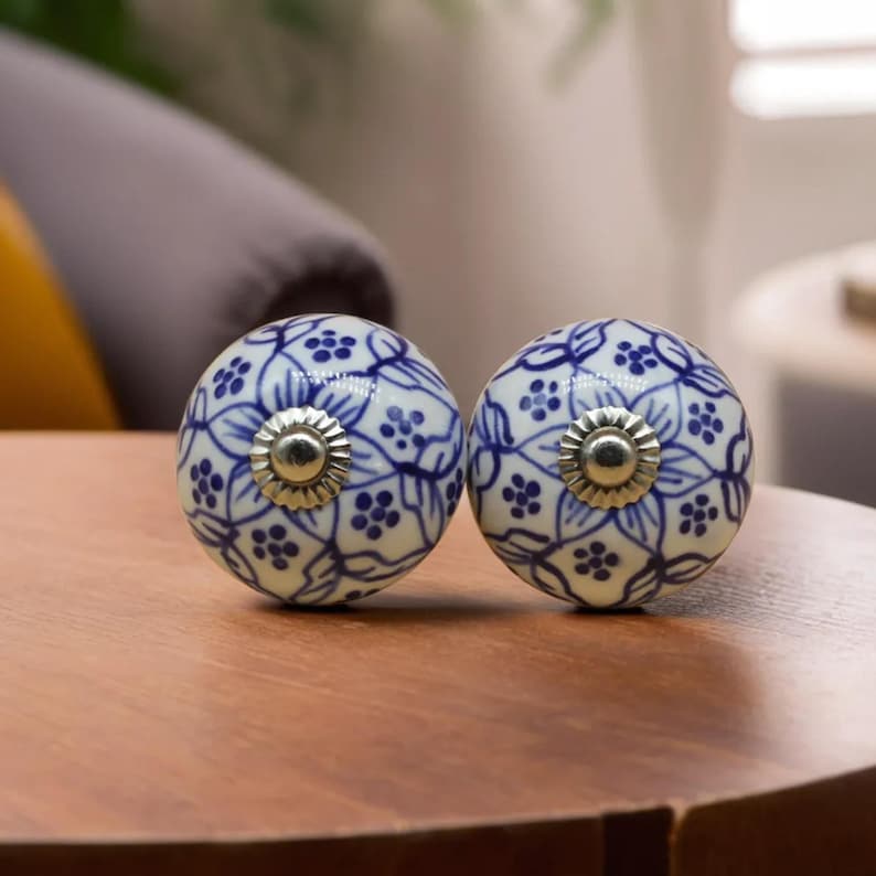 May include: Two round, white ceramic cabinet knobs with a blue floral pattern. Each knob has a silver-colored metal base. The knobs are on a wooden surface.