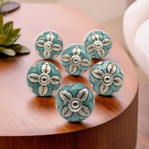 Może przedstawiać: Sześć turkusowych ceramicznych gałek do szafek z kwiatowym wzorem. Każda gałka ma srebrny środek i wzór białych i turkusowych płatków kwiatów. Te dekoracyjne gałki są idealne, aby dodać odrobinę elegancji do każdego mebla.