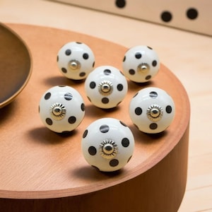 Könnte beinhalten: Sechs weiße Keramik-Schrankknöpfe mit schwarzen Polka-Dots. Jeder Knopf hat einen silberfarbenen Sockel und eine dekorative Mitte. Die Knöpfe sind auf einer Holzoberfläche angeordnet, um ihr Design und ihren Einsatz in der Inneneinrichtung zu zeigen.