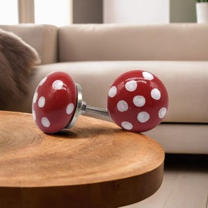Könnte beinhalten: Zwei rote, pilzförmige Schrankknöpfe mit weißen Tupfen. Die Knöpfe sind an einer Metallschraube und -scheibe befestigt. Die Knöpfe liegen auf einem Holztisch. Wohndekoration.