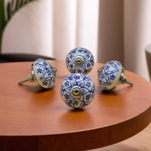 May include: Four white and blue ceramic cabinet knobs with a floral design are arranged on a wooden surface. Each knob has a silver metal screw and a decorative center. The knobs are a decorative home decor item.