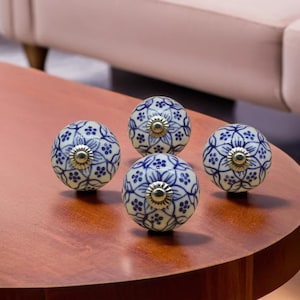 May include: Four ceramic cabinet knobs with a blue and white floral design. Each knob has a brass-colored metal base. The knobs are spherical and rest on a wooden surface.