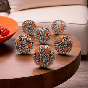 May include: Six orange ceramic cabinet knobs with a floral design in white and gray are arranged on a wooden table. The knobs are round and have a glossy finish. An orange bowl with red berries is in the background.