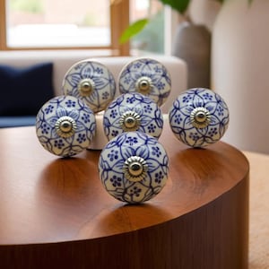 May include: A collection of six ceramic cabinet knobs. Each knob is round, featuring a white base with a blue floral pattern and a silver-colored metal center. The knobs are arranged on a wooden surface.