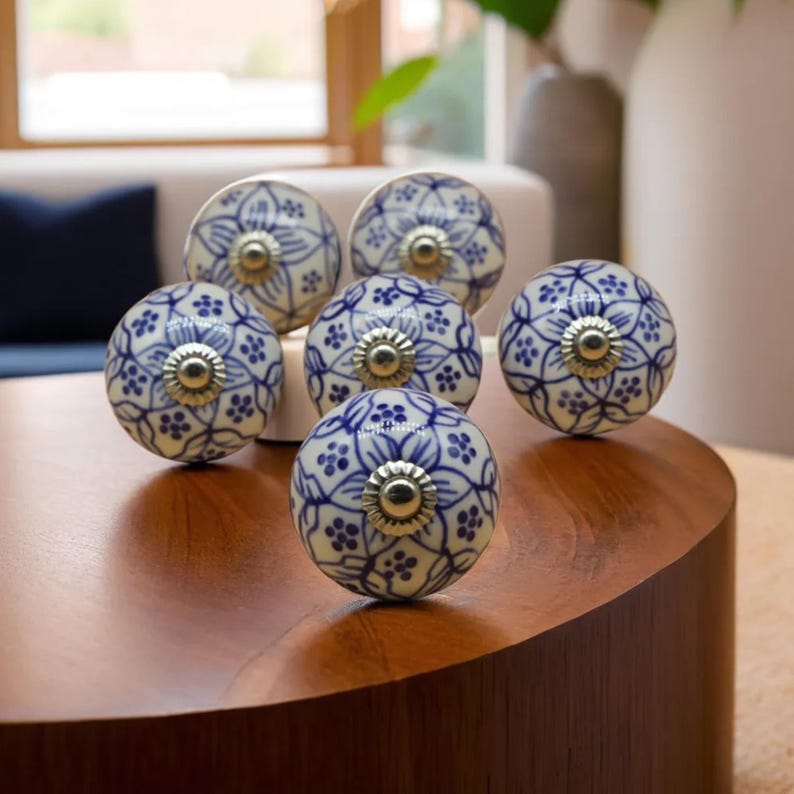 May include: A collection of six ceramic cabinet knobs. Each knob is round, featuring a white base with a blue floral pattern and a silver-colored metal center. The knobs are arranged on a wooden surface.
