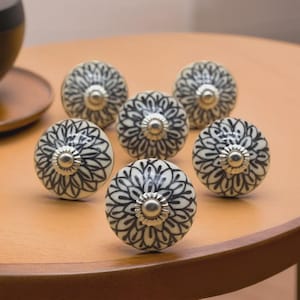 May include: Six decorative ceramic cabinet knobs. Each knob is off-white with a black floral design and a silver-colored metal center. The knobs are arranged on a light brown wooden surface.