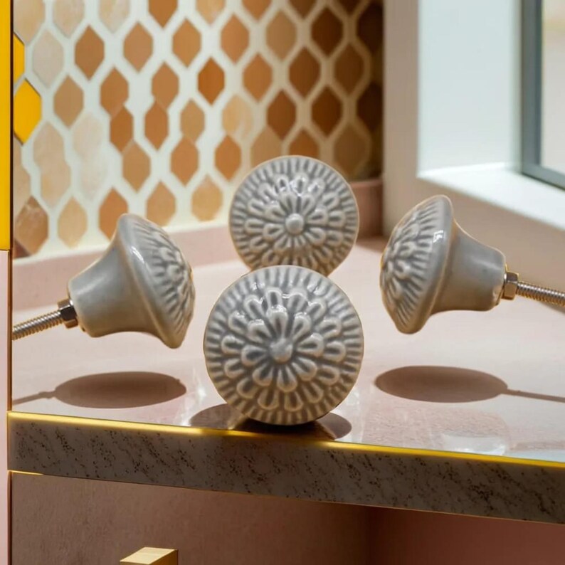 Peut inclure: Quatre boutons de meuble en c&eacute;ramique grise avec un motif floral. Les boutons sont ronds avec une base l&eacute;g&egrave;rement conique et sont mont&eacute;s sur des vis argent&eacute;es. Les boutons sont dispos&eacute;s sur une surface rose avec une bordure dor&eacute;e, sur fond de carrelage &agrave; motifs.