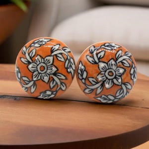 May include: Two orange ceramic cabinet knobs with white floral and leaf designs. The knobs have a glossy finish and are set on a wooden surface. The floral pattern is centered on each knob.