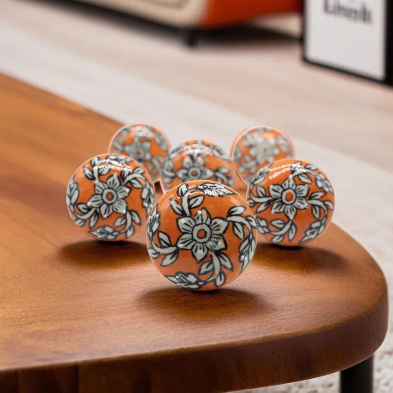 May include: Six orange ceramic cabinet knobs with a floral design in white and gray. The knobs are round and have a glossy finish. The knobs are arranged on a wooden surface.