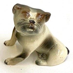 May include: A ceramic bulldog figurine in a seated position. The dog has a cream-colored body with brown patches on its head and back. The figurine has a detailed face with a wrinkled snout and small, dark eyes. The dog's ears are brown, and its paws are extended.