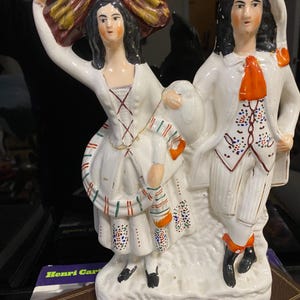 Puede incluir: Una figura de porcelana vintage que representa a una pareja con vestimenta de época. La mujer viste un vestido blanco con detalles coloridos, y el hombre lleva un traje blanco con una corbata naranja. La figura tiene un diseño detallado pintado a mano.