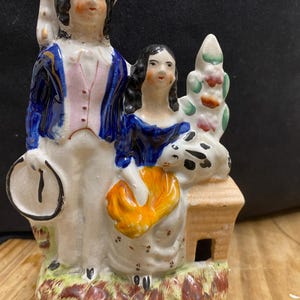 Op de afbeelding: Een vintage keramieken beeldje dat een man en een vrouw in kleding uit die tijd afbeeldt. De man draagt een blauwe jas en houdt een klok vast. De vrouw zit en houdt een geel voorwerp vast. De basis heeft een kleine structuur en geschilderde bloemendetails.