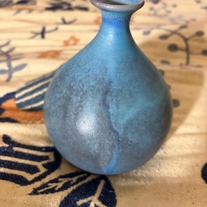 May include: A small, round ceramic vase with a narrow neck and a flared rim. The surface features a mottled glaze with varying shades of blue, transitioning from a bright, vibrant sky blue at the top to a deeper, darker, and more muted navy blue.