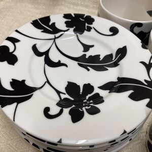 Könnte beinhalten: Ein Stapel weißer Keramikteller mit einem schwarzen Blumenmuster. Die Teller gehören zu einem Geschirrset. Weitere Teile des Sets, darunter Schalen und ein kleiner Krug, sind ebenfalls zu sehen. Glänzende Oberfläche.