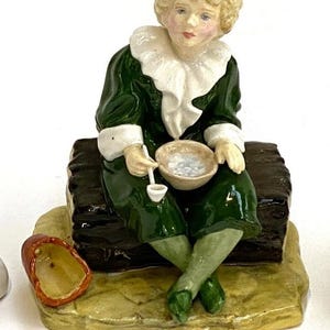 Puede incluir: Una figura de porcelana de una figura sentada con un abrigo verde con ribete blanco y cuello con volantes. La figura sostiene un cuenco y una pipa. La figura está sobre una base marrón con una base beige y verde.