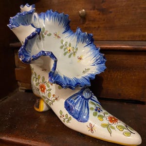 May include: A white ceramic shoe-shaped vase with blue ruffled trim and floral designs. The vase features hand-painted flowers in red, yellow, and blue, with green leaves. The heel is yellow. A decorative piece for home decor.