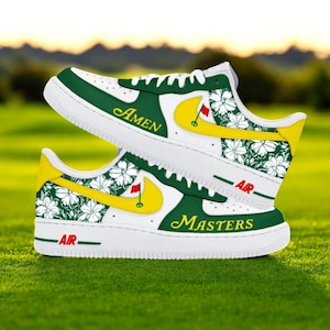 Peut inclure: Baskets blanches avec des détails verts et jaunes. Le design comprend des motifs floraux, le mot "Amen" et un drapeau de golf. Le mot "Masters" est également visible. Les chaussures sont sur une surface herbeuse verte.