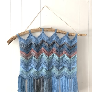 May include: A blue and gray crocheted wall hanging with a fringe border. The hanging is attached to a piece of driftwood with twine.