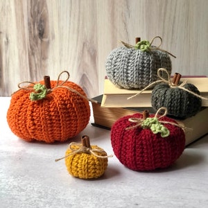 May include: Four crocheted pumpkins in different sizes and colors. The pumpkins are orange, red, gray, and yellow. They are arranged on a white surface with a stack of books in the background.