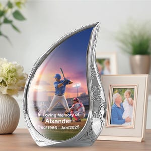 May include: A silver teardrop-shaped memorial with a baseball scene inside. The text "In Loving Memory of Alexander Dec/1956 - Jan/2025" is visible. A framed photo is in the background.