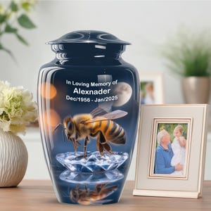May include: A navy blue cremation urn with a bee design and the text "In Loving Memory of Alexnader Dec/1956 - Jan/2025". The urn features a detailed illustration of a bee and a diamond-shaped base. A framed photo is next to the urn.