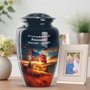 May include: A black cremation urn with a glossy finish, depicting a sunset scene with an American flag. The urn is inscribed with "In Loving Memory of Alexnader Dec/1956 - Jan/2025." A framed photo is next to the urn.