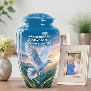 May include: A blue cremation urn featuring a dove design and the inscription "In Loving Memory of Alexnader Dec/1956 - Jan/2025." A framed photograph of a man and child sits beside the urn. The urn is on a wooden surface.