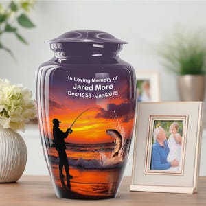 May include: Purple cremation urn with a fishing scene at sunset. The urn has a glossy finish and features the text "In Loving Memory of Jared More Dec/1956 - Jan/2025". A framed photo sits beside the urn.