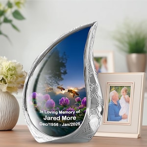 May include: A silver memorial keepsake with a teardrop shape. The interior features a scenic image of bees, purple flowers, and a blue sky. Text reads "In Loving Memory of Jared More Dec/1956 - Jan/2025". A framed photo is in the background.