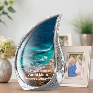 May include: A silver-toned memorial urn in a teardrop shape, featuring a scenic ocean view. The urn is engraved with the text "In Loving Memory of Jared More Dec/1956 - Jan/2025". A framed photo is visible in the background.