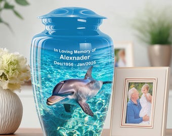 May include: A blue cremation urn with a dolphin design. The urn has a glossy finish and features the text "In Loving Memory of Alexnader Dec/1956-Jan/2025". A framed photo is next to the urn.